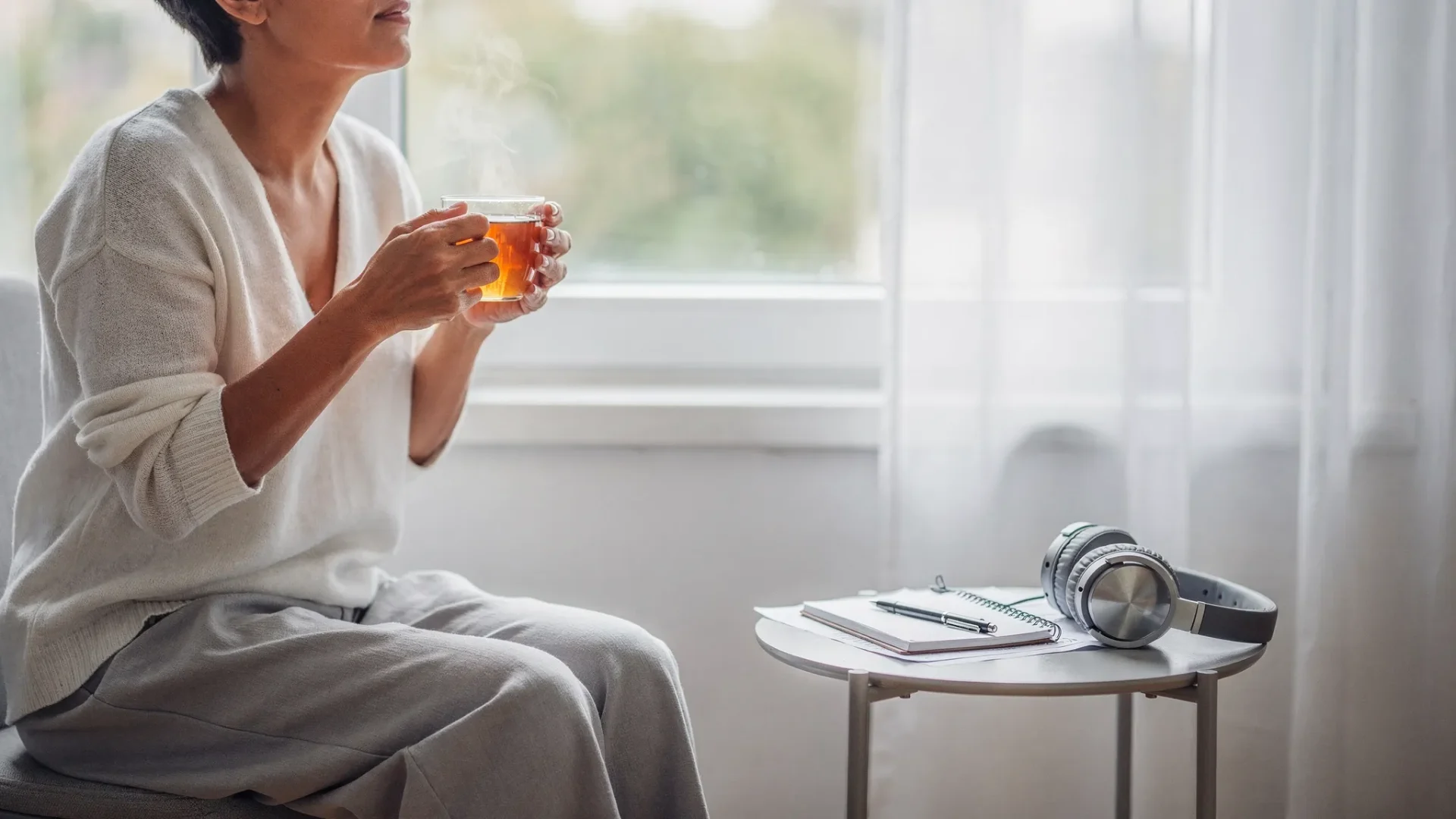 Eine ruhige, helle Szene: Eine erwachsene Person sitzt mit einer Tasse Tee am Fenster auf einem Stuhl, atmet sichtbar tief durch. Auf dem Beistelltisch liegen Kopfhörer und ein Notizbuch mit Stift. Die Stimmung vermittelt Entlastung und sofortige Unterstützung.