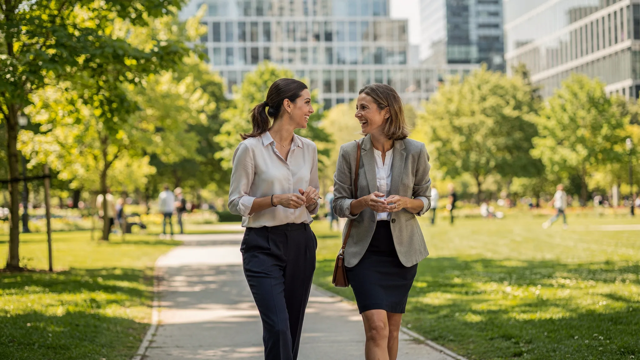 Zwei Kolleginnen gehen in der Mittagspause durch einen grünen Stadtpark, sprechen miteinander und genießen helles Tageslicht; im Hintergrund moderne Bürogebäude.